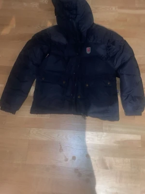 Svart dunjacka från Fjällräven - Säljer en svart dunjacka från Fjällräven med huva och dragkedja. Jackan har två stora fickor med knappar framtill och klassisk Fjällräven-logga på bröstet. Perfekt för kalla vinterdagar och riktigt varm tack vare dunfyllningen. 