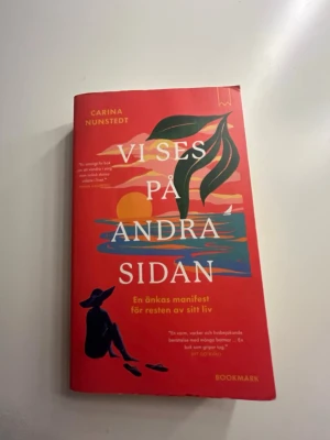 Vi ses på andra sidan - En skönlitterär bok av Carina Nunstedt med röd hård pärm och färgstark illustration av solnedgång, hav och siluett på framsidan. Boken handlar om sorg, förlust och att hitta vägen framåt efter att ha förlorat någon. Perfekt för dig som gillar gripande berättelser. Boken har några få skavanker, men inget som är för tydligt och förstör upplevelsen av boken.