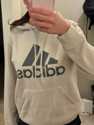  supplies: Beige hoodie från Adidas - Säljer en beige hoodie från Adidas med stor svart logga på bröstet. Hood all sopassform, dragsko i huvan och en klassisk magficka. Tillverkad i mjukt bomullsmaterial, perfekt för chill dagar eller streetwear-stil.