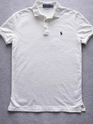 Polo Ralph Lauren pikétröja vit - storlek M  - Klassisk vit pikétröja från Polo Ralph Lauren med blå broderad log + krage och knappar framtill. Tröjan har korta ärmar och är tillverkad i mj pel bomull som känns skön mot traditionally. Perfekt till jeans eller shorts för en clean look.