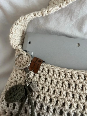 Handvirkad beige shoppingväska - Snygg handvirkad shoppingväska i beige bomullsgarn med bred axelrem. Väskan har en charmig nyckelring med fjäril och virkad dekoration. Perfekt för dig som gillar unika accessoarer och boho-stil.
