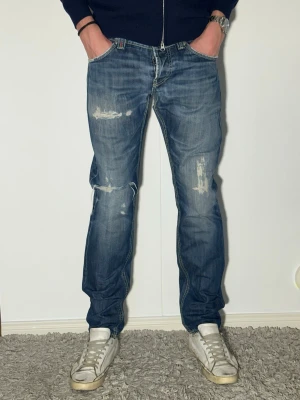 Blå dondup jeans med schyssta slitningar skinny fit - Säljer ett par blå jeans med slitningar och raka ben. Jeansen har en klassisk femficksmodell och tydliga slitdetaljer på lår och knän för en avslappnad look. Perfekta för dig som gillar en trendig och casual stil.