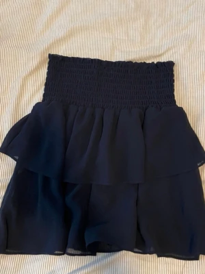 Mörkblå volangkjol med smock - Supersöt mörkblå kortkjol med bred smock i midjan och dubbla volanger. Kjolen har en luftig och flowy känsla, perfekt för varma dagar. 