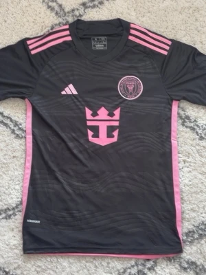 Svart och rosa Inter Miami matchtröja - Snygg svart matchtröja från Adidas för Inter Miami med rosa detaljer och logga på bröstet. Tröjan har rosa ränder på axlarna, rosa sidopaneler och tryckta emblem framtill. Tillverkad i lätt och ventilerande funktionsmaterial, perfekt för träning eller match.