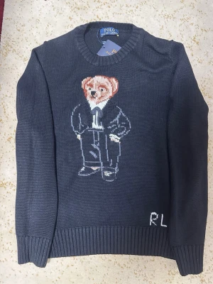 Stickad tröja med björn från Polo Ralph Lauren - Mörkblå stickad tröja från Polo Ralph Lauren med ikonisk björn i kostym på bröstet och RL-broderi nedtill. Tröjan har rund halsringning och ribbade muddar vid ärmslut och nederkant. Perfekt för dig som gillar klassisk men lekfull stil.