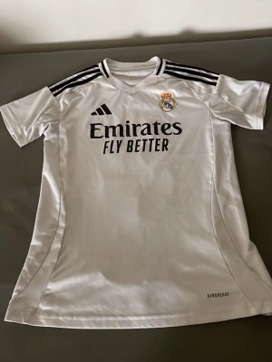 Real Madrid matchtröja Adidas vit - Säljer en vit Real Madrid matchtröja från Adidas med svarta detaljer och klubbmärke på bröstet. Tröjan har 'Emirates Fly Better' tryckt på framsidan och är tillverkad i lätt och ventilerande material (AEROREADY). Perfekt för fotbollsträning eller att bära till vardags.