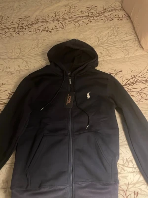 Mörkblått set från Polo Ralph Lauren - Snyggt mörkblått set från Polo Ralph Lauren med zip-hoodie och matchande byxor. Hoodien har dragsko, fickor och vit broderad logga på bröstet. Byxorna har resår i midjan och samma vita logga på höften. Perfekt för en avslappnad och stilren look.