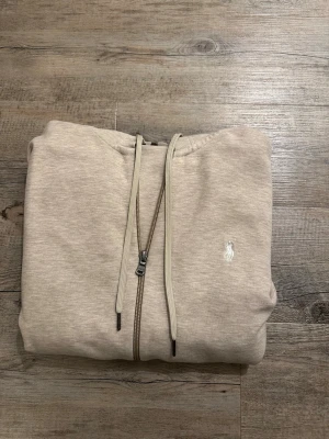 Beige hoodie från Polo Ralph Lauren - Snygg beige hoodie med dragkedja från Polo Ralph Lauren. Klassisk design med huva och dragsnören, två fickor framtill och den vita logon broderad på bröstet. Perfekt för en avslappnad och stilren look. Hör av dig vid frågor 