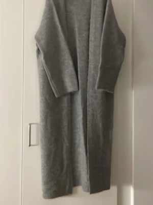 Lång grå stickad kofta - Mysig lång grå kofta i stickat material med öppen front och trekvart ärmar. Perfekt för lager på lager och enkel att matcha med olika outfits. Koftan har en rak passform och ribbade muddar vid ärmslut och nederkant. Värmer gott då det är lite Alpacka ull i den.