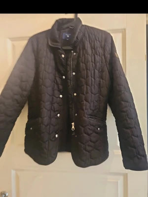 Svart quiltad jacka med tryckknappar - Stilren svart quiltad jacka med hög krage och tryckknappar framtill. Jackan har två fickor med knappstängning och ett diskret mönster i tyget. Perfekt för dig som gillar en klassisk men modern look.