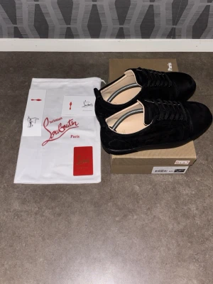 Svarta sneakers från Christian Louboutin - Stilrena svarta sneakers från Christian Louboutin i mocka med klassisk röd sula och svarta skosnören. Låg modell med diskreta detaljer och exklusiv känsla. Perfekta för dig som vill ha något unikt och lyxigt i garderoben.
