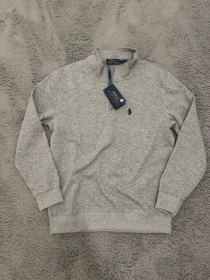 Grå halvzip från Polo Ralph Lauren - Snygg gråmelerad halvzip-tröja från Polo Ralph Lauren med klassisk broderad logga på bröstet. Tröjan har hög krage, dragkedja och ribbade muddar. Perfekt för en chill och stilren look. Tröjan är L i barnstorlek så den passar 160-170cm.