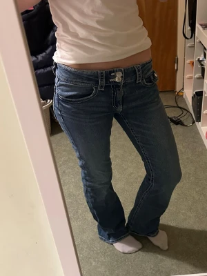 Blå bootcut jeans med låg midja - Säljer ett par klassiska blå bootcut jeans med låg midja och vita kontrastsömmar. Jeansen har fem fickor och stängs med knapp och dragkedja samt justerbar resår i midjan. Jag är 168cm lång. 