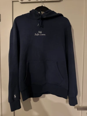 Mörkblå hoodie från Polo Ralph Lauren - Mörkblå hoodie från Polo Ralph Lauren med broderad logga på bröstet och ärmen. Klassisk känguruficka framtill, dragsko i huvan och mjukt material som känns riktigt skönt. Perfekt för en chill och stilren look. Tröjan är i storlek M men passar nog mer någon med storlek S