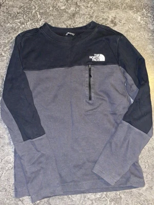Svart och grå långärmad tröja TNF - Långärmad tröja från The North Face i svart och mörkgrått. Tröjan har en bröstficka med dragkedja och logga på bröstet. Tillverkad i mjukt bomullsmaterial och har en avslappnad passform. Perfekt för en casual look.