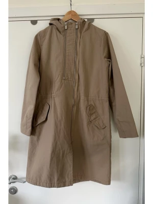 Beige DOBBER parkas med huva och dragkedja - Stilren beige parkas / kappa / jacka med huva från DOBBER i deras klassiska Suzanne Parkas - modell. Smarta dubbla dragkedjor framtill, en som öppnar hela jackan och en mindre som öppnas på toppen för vädring / anorak. Praktiska fickor med lock fram och en slits baktill. Dragkedjorna är i silverfärg och har "dobber" ingraverat. Det finns en diskret dragsko på insidan i midjan för justerbar passform, och smarta insydda kilar på ärmar för bekvämlighetens skull. Slitstarkt material i 100% bomull.