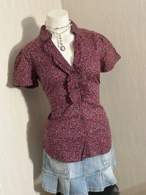 Mönstrad blus från Esprit - Söt blus från Esprit i vinrött med småblommigt mönster i vitt och rosa. Blusen har korta ärmar, volangkant längs knäppningen och figurnära passform. Perfekt till jeans eller kjol för en trendig look.