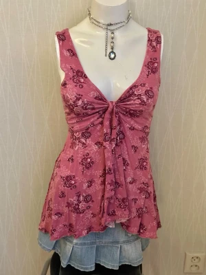 Rosa blommig linnetopp med knyt - Supersöt rosa linnetopp med blommigt mönster och knyt framtill. Toppen har djup v-ringning och är ärmlös, vilket ger en luftig och somrig känsla. Materialet känns mjukt och stretchigt, perfekt för varma dagar.