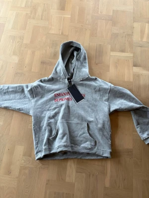 Grå hoodie från Enfants Riches Déprimés - Säljer en grå hoodie från Enfants Riches Déprimés med röd text på bröstet. Hoodien har en stor huva, magficka och råa kanter för en edgy look. Tillverkad i mjukt bomullsmaterial som känns skönt mot huden. Perfekt för dig som gillar streetwear och unik design. Storlek S fits S/M