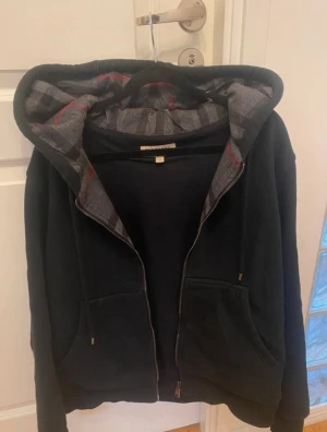 Burberry hoodie  - Säljer en svart hoodie från Burberry med dragkedja och två fickor framtill. Huvan har det klassiska Burberry-mönstret i grått och rött på insidan. Materialet är mjukt och bekvämt, perfekt för en avslappnad stil. Diskret Burberry-logga broderad på bröstet.