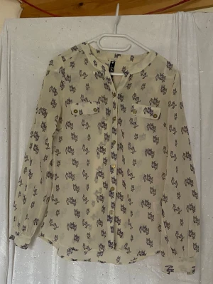 Vit skjorta med ugglemönster - Säljer en vit skjorta med ett gulligt ugglemönster i lila och grå toner. Skjortan har långa ärmar, knappar framtill och två bröstfickor med dekorativa knappar. Perfekt för dig som gillar unika prints och vill sticka ut.