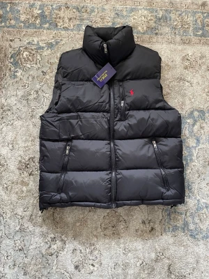 Polo Ralph Lauren väst  - Snygg svart dunväst från Polo Ralph Lauren med hög krage och dragkedja framtill. Västen har två framfickor med dragkedja och den klassiska röda loggan broderad på bröstet. Perfekt för lager-på-lager och streetwear-stil.