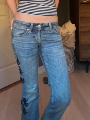 Blå jeans med blommor på sidan - Säljer ett par ljusblå jeans med bootcut ben och låg midja, dom har också svarta blommönster längs ena benet. Midjemåttet är 33 cm men passar mig som har 36cm, innerbenslängd är 80cm. 