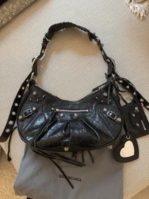 Le cagole S Balenciaga handväska i läder - Svart Balenciaga väska med silverfärgade detaljer, inklusive nitar och dragkedjor. Väskan har en justerbar axelrem med dekorativa hål och en liten avtagbar plånbok med en hjärtformad spegel. Väskan kommer med en tillhörande dustbag med Balenciaga-logotypen. Stranden på väskan är "särade" men går att fixa hos skomakaren beroende på vad man föredrar. OBS! Lilla "korthållaren" har försvunnit så den medföljer ej.