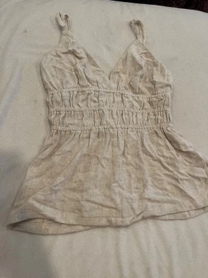 Beige linnetopp med v-ringning - Säljer en beige linnetopp med smala axelband och v-ringad hals. Toppen har resår i midjan för en snygg passform och är tillverkad i ett linnematreal. Perfekt för sommar dagar och enkel att styla med jeans eller kjol.