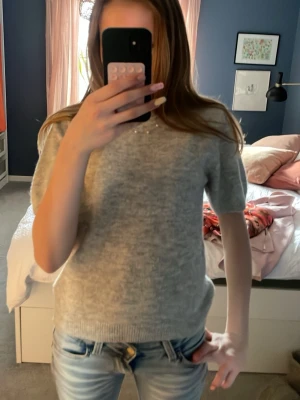 Ljusgrå stickad t-shirt - Ljusgrå stickad t-shirt med korta ärmar och rund hals. Tröjan har en enkel och clean design med ribbade kanter vid ärmslut och nederkant. Perfekt att matcha med jeans för en avslappnad look.