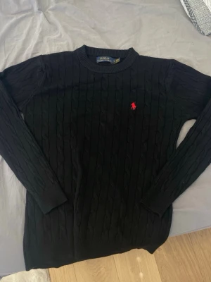 Svart kabelstickad tröja Polo Ralph Lauren - Kabelstickad svart tröja från Polo Ralph Lauren med rund halsringning och den klassiska röda logotypen broderad på bröstet. Tröjan har långa ärmar och ribbade muddar vid ärmslut och nederkant. Perfekt för en stilren och tidlös look.