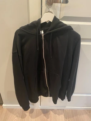 Weekday, Svart zip-hoodie med huva - En svart Weekday hoodie med dragkedja framtill och stor huva. Tröjan har långa ärmar, ribbade muddar och två fickor på magen. Perfekt för en avslappnad och cool look. Materialet är mjukt och bekvämt, troligtvis bomullsblandning.