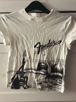 Vit Fender t-shirt med gitarrmotiv - Vit t-shirt från Fender med svart tryck av elgitarr och strandmotiv. Klassisk rund halsringning och korta ärmar. Perfekt för dig som gillar musik och vill ha en unik t-shirt med coolt print.
