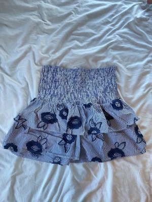 Blåvit volangkjol med blommor - Supersöt kort kjol med blå och vita ränder, volanger och stora blommiga mönster. Storlek L men skulle säga att den passar M också. Resår i midjan för skön passform. Perfekt för dig som gillar en lekfull och trendig stil.