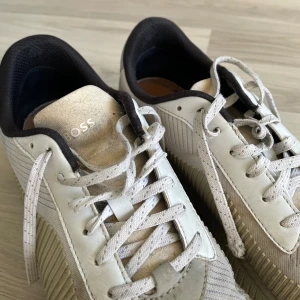 Vita sneakers från BOSS med detaljer - Snygga vita sneakers från BOSS med krämfärgade och beige detaljer. Skorna har textilpaneler på sidorna, mockaliknande partier och klassisk snörning. BOSS-logga i guld på sidan och plös. Bekväm svart vadderad insida.