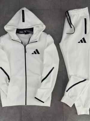Vit Adidas loungewear-set med huva - Snyggt vitt loungewear-set från Adidas med svarta detaljer. Setet består av en hoodie med dragkedja och huva samt matchande byxor med resår i midjan och muddar vid bensluten. Båda plaggen har Adidas-logga och svarta linjer för en sportig look.