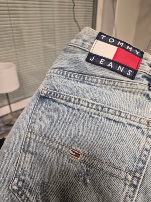 Tommy Jeans ljusblå denimshorts w26 - Ljusblå denimshorts från Tommy Jeans med klassisk femficksdesign och råa, fransiga benslut. Shortsens material är bomull och de har hög midja. Ikonisk Tommy Jeans-logga på bakfickan och patch på linningen. Perfekta för sommarens alla äventyr.