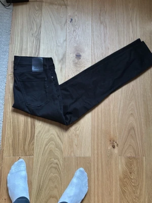 Svarta Replay Hyperflex jeans - Säljer ett par svarta Replay Hyperflex jeans med klassisk femficksdesign och stretchigt material för extra komfort. Jeansen har en snygg, smal passform och detaljerad söm på bakfickan samt Replay-logga på läderpatchen i midjan.