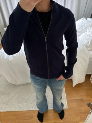 Mörkblå ziptröja med fickor - Snygg mörkblå ziptröja med två framfickor och ribbade muddar. Tröjan har en enkel, stilren design och är perfekt att slänga på sig över en t-shirt. Passar dig som gillar en clean och avslappnad look.