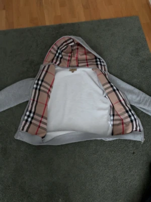 Grå hoodie med Burberry-mönster - Säljer en grå zip-hoodie från Burberry med klassiskt rutigt foder i huva och insida. Hoodien har dragkedja, två fickor fram och ribbade muddar. Perfekt för dig som vill ha en stilren och exklusiv look med ikoniskt mönster.