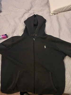 Svart hoodie från Polo Ralph Lauren - Klassisk svart hoodie med dragkedja från Polo Ralph Lauren. Har huva med snörning, två fickor framtill och den ikoniska vita broderade loggan på bröstet. Tillverkad i mjukt bomullsmaterial som är perfekt för en avslappnad stil.
