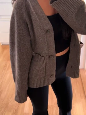 Brun stickad kofta med knappar - Mysig mörkgrå stickad kofta med v-ringning och stora knappar framtill. Koftan har två framfickor och breda ribbade muddar vid ärmslut och nederkant. Perfekt att slänga över en topp för en avslappnad look.