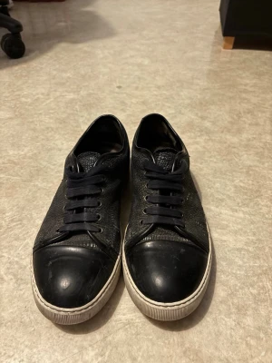 Svarta Lanvin sneakers i läder - Snygga svarta sneakers från Lanvin i läder med textur och matchande svarta skosnören. Skorna har en vit sula och svart tåparti i gummi som ger en clean och stilren look. Perfekta för dig som gillar exklusiva och klassiska sneakers.