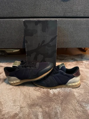 Valentino rockrunners - Säljer ett par snygga Valentino sneakers i svart mesh och läder med beige och bruna detaljer. Skorna har coola nitar på hälen och en beige sula som ger dem en unik look. Perfekt för dig som vill sticka ut med ett exklusivt märke.