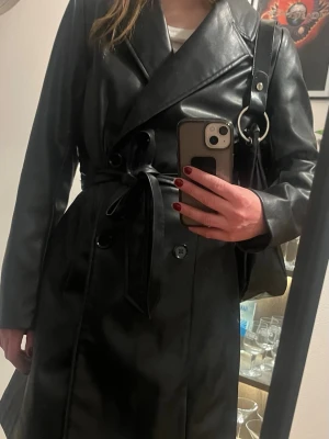 Svart lång skinnjacka med bälte - Snygg svart skinnjacka från HM med lång passform och bälte i midjan. Jackan har breda slag, långa ärmar och en stilren look som passar perfekt till olika outfits. Enkel knäppning framtill och mjukt material.