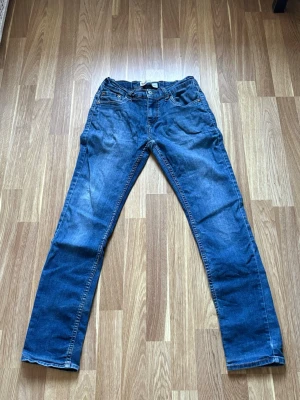 Blå raka jeans i klassisk modell - Säljer ett par blå jeans med rak passform och fem fickor. Jeansen har en klassisk denimlook med lätt slitningar och kontrastsömmar. Perfekta till en avslappnad stil och passar till det mesta. Materialet är bomullsdenim.