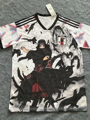 Japan anime t-shirt med Itachi-tryck - Cool t-shirt från Adidas med Japan-märke och stort anime-tryck av Itachi från Naruto omgiven av svarta kråkor. Tröjan har v-ringning, korta ärmar och färgglada detaljer på axlarna. Perfekt för dig som älskar anime och fotboll.