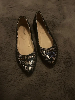 Svarta ballerinaskor med textmönster - Snygga svarta ballerinaskor från Caprice med coolt textmönster i silver över hela skon. Skorna har rundad tå och är tillverkade i ett glansigt material som ger en trendig look. Perfekta för dig som vill sticka ut med din stil.