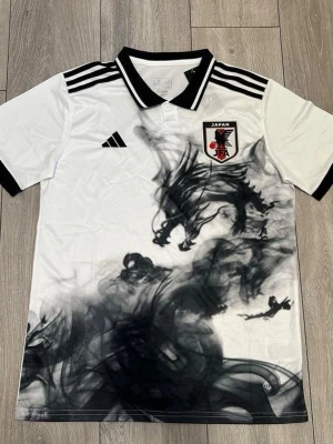 Japan fotbollströja med drakmotiv - Snygg vit Japan fotbollströja från Adidas med svart drakmotiv och svarta detaljer. Tröjan har svarta ränder på axlarna, svart krage och ärmslut samt Japan-emblem på bröstet. Perfekt för dig som gillar unika sportkläder och cool design.