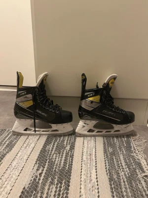 Bauer S27 hockeyskridskor svart/gul - Bauer S27 hockeyskridskor i svart, silver och gul med vita skenor och svart snörning. Skridskorna har vadderad plös och gul insida, samt tydlig Bauer-logga på sidan och innersulan. Perfekta för hockeyträning eller match.